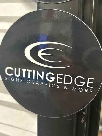 Round black sign with 'Cutting Edge Signs Graphics & More' logo on a reflective surface.