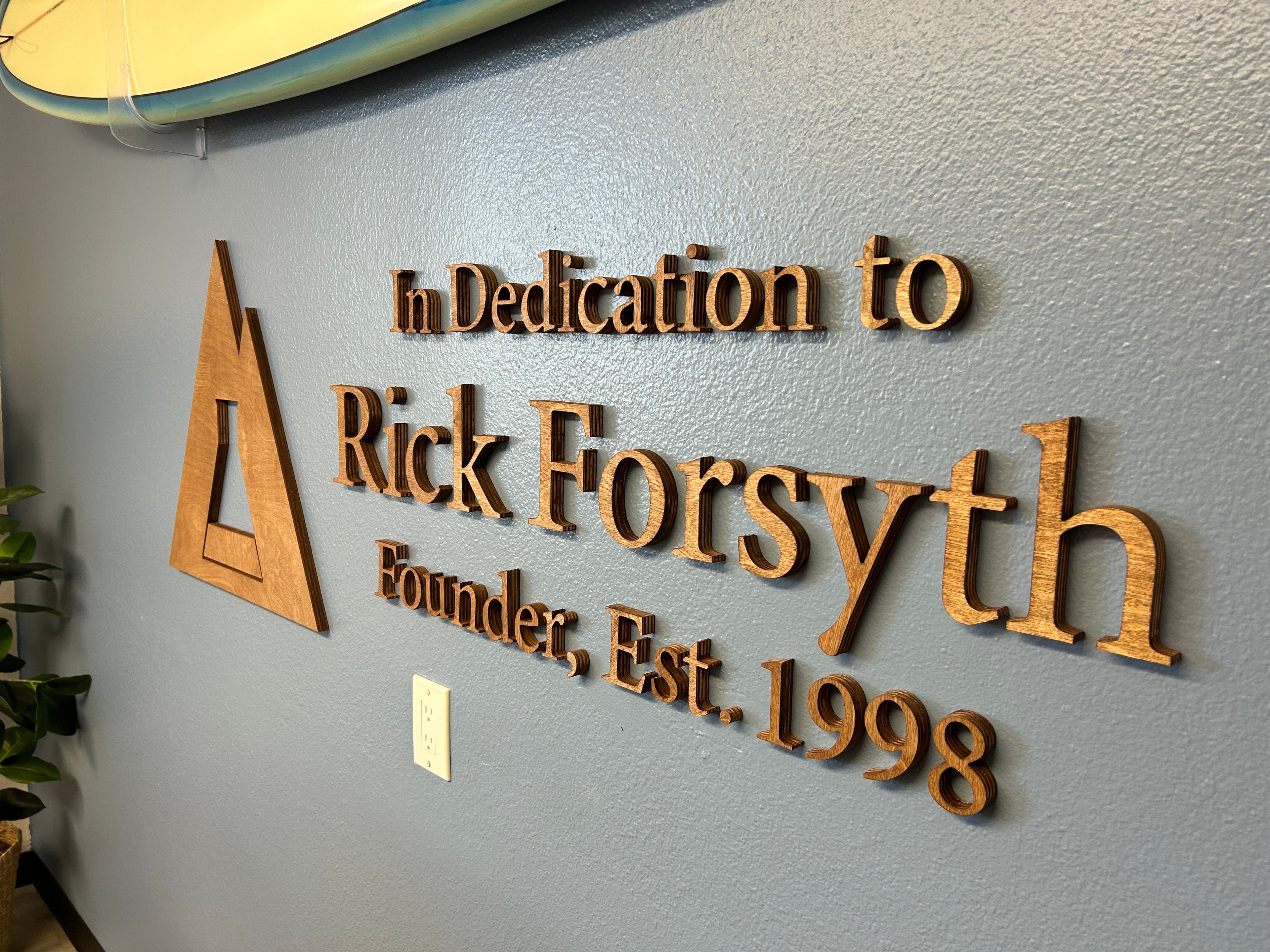 Wooden plaque on a wall with text 'In Dedication to Rick Forsyth, Founder, Est. 1998' on a gray background.