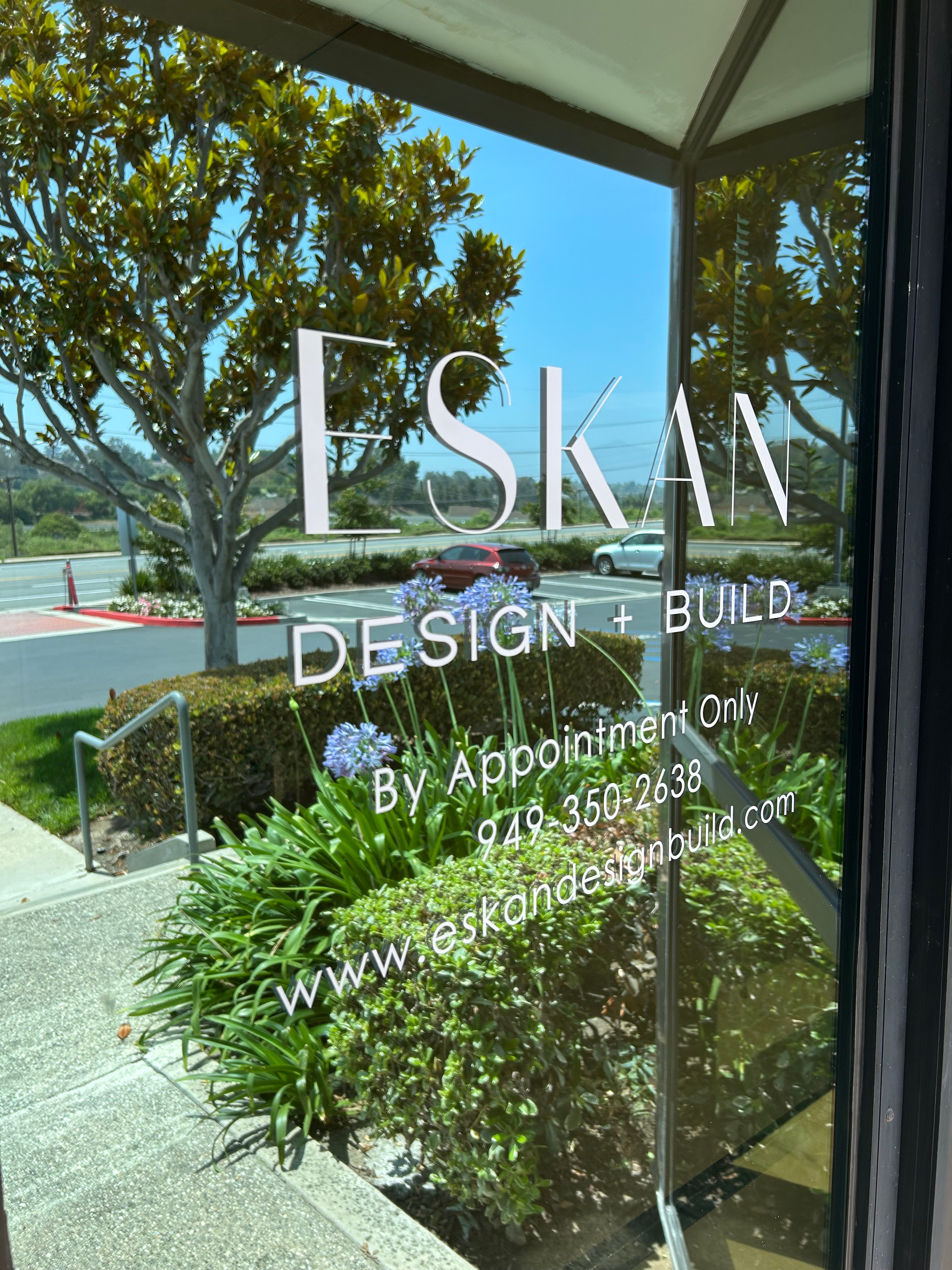 Glass window with 'ESKAN' design and build branding, reflecting trees and sky.